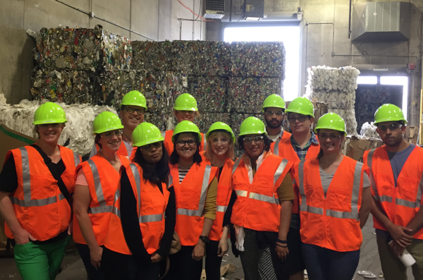 Students in hard hats and orange safety vests stand in front of 20-foot bales of paper to be recycled