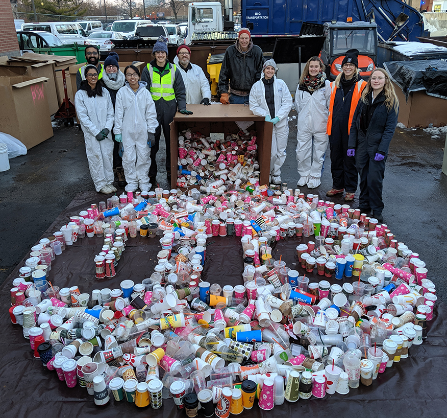 waste audit volunteer stand over thousands of disposed single-use beverage cups designed to look like the UIC logo.