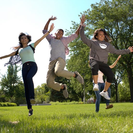 UIC students jumping and smiling on a grassy area on campus.