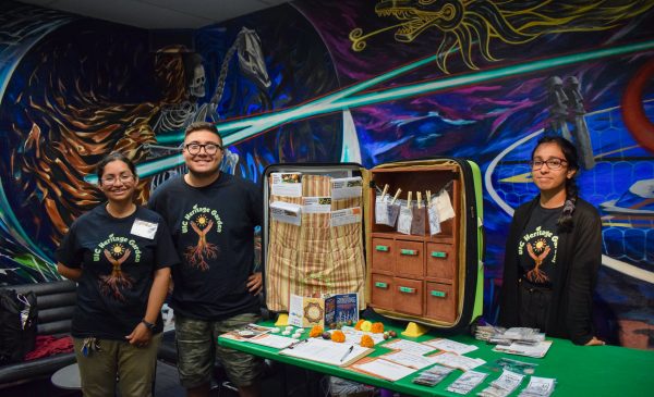three members of the UIC Heritage Garden stand smiling in front of an open suitcase that contains seeds and educational print-outs.